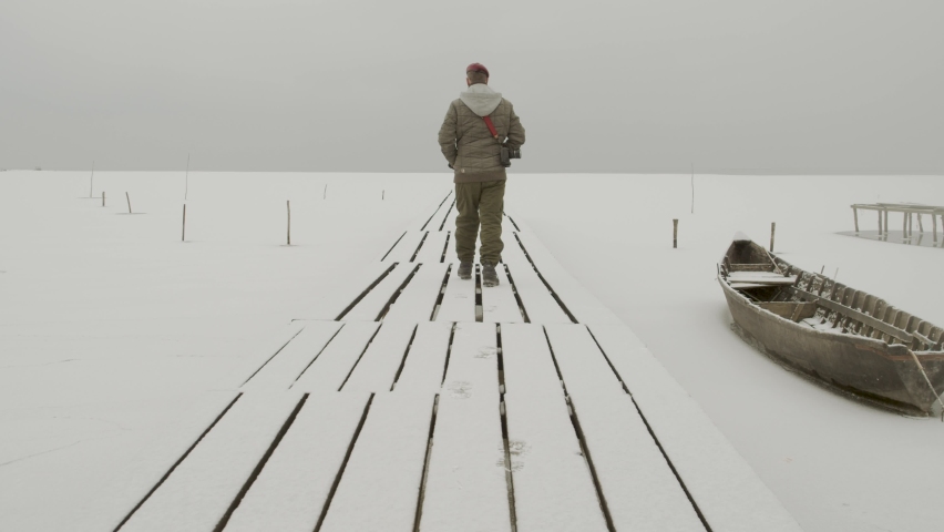 lonely man walking on snowy wooden bridge and fishing boats at shore