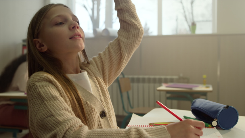 Cheerful girl studying in classroom. Cute schoolgirl writing in notebook. Portrait of smart pupil raising hand at lesson. Female student answering on question in school class 
