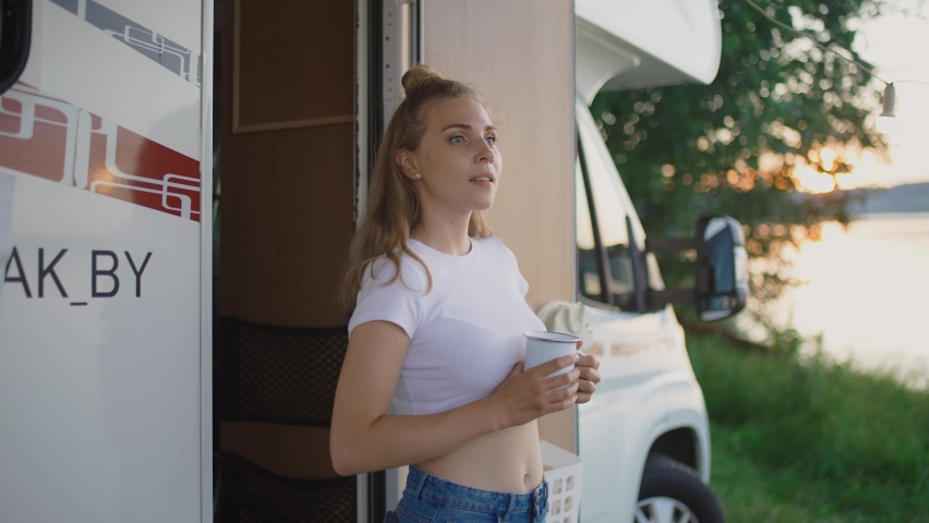 Caucasian female is resting in nature near the lake, standing near a mobile home and enjoying nature holding a mug of tea in her hands, summer vacation, woman near camping car, 4k slow motion.