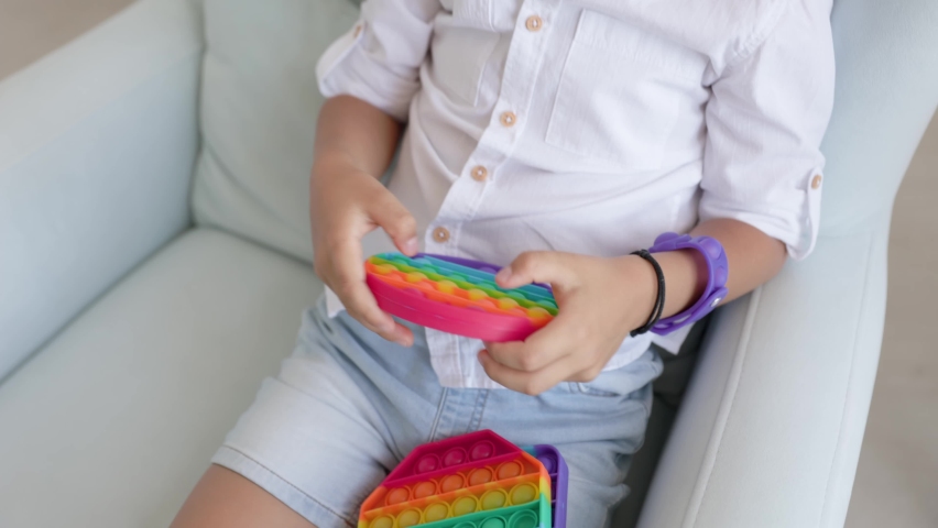 Portrait blonde boy with pop it sensory toy different forms. Boy presses colorful rainbow squishy soft silicone bubbles in children