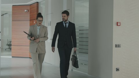 Medium PAN slowmo shot of couple of young confident lawyers in formalwear having conversation about work while walking along modern office