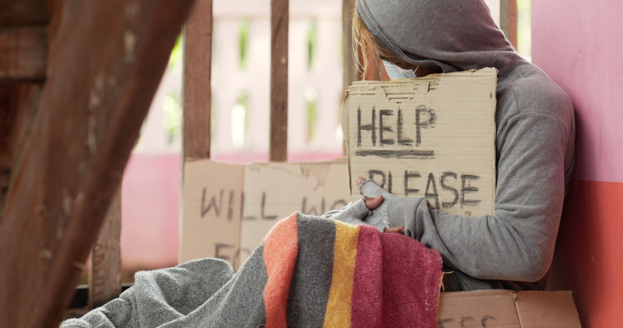 Closeup of Refugee homeless man in face mask hug help please label. Tired and sad male outdoor. Young man sitting alone. Vagrant person begging for money.