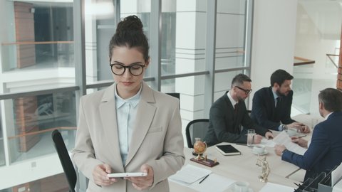 PAN medium slowmo portrait of young female lawyer in eyeglasses and formalwear posing for camera with digital tablet in hands while her colleagues having discussion at meeting table
