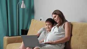 Pregnant woman and boy talking by video call on laptop while sitting on couch, slow motion.