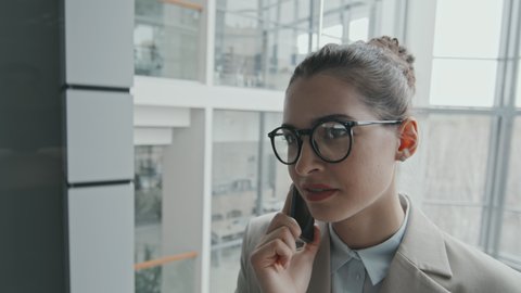 Slow-motion close-up of young brunette businesswoman or lawyer in eyeglasses and formalwear talking on smartphone while going down in glass elevator in office