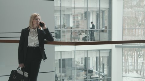 Slow-motion wide medium PAN shot of successful mid-adult female lawyer in formalwear talking on smartphone leaning on railings in modern office