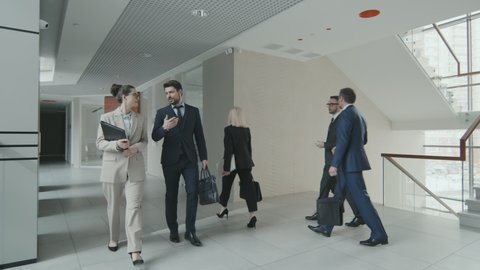 Slowmo shot of male and female lawyers or business people in suits and formalwear talking while walking along modern office working together in law firm
