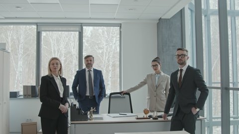 Medium slowmo portrait of confident law firm founder in suit posing for camera with team of professional male and female lawyers standing together by table in office room