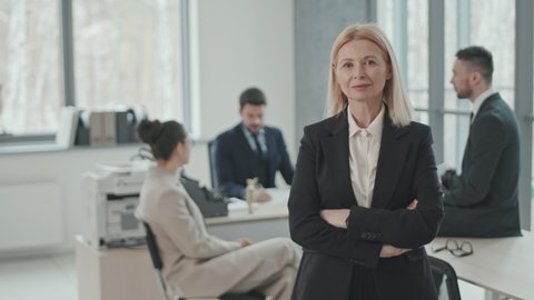 PAN medium slowmo of successful mid-adult female lawyer in formalwear smiling to camera standing with hands folded in modern office room while colleagues discussing case in background