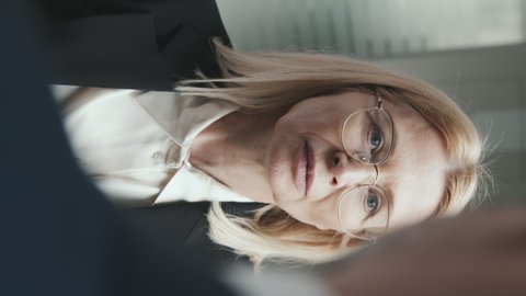Vertical medium close-up of mid adult blonde female lawyer in eyeglasses and formalwear listening to businessman in office