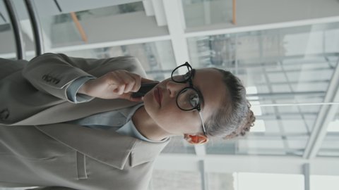 Vertical medium slowmo shot of young female lawyer in eyeglasses and formalwear making phone call while going down in glass elevator in office
