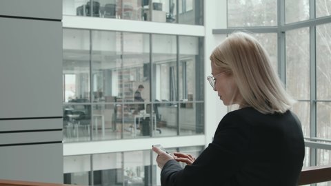 Rear-view slowmo shot of mid adult blonde businesswoman in formalwear using smartphone while leaning on railing in contemporary glass office