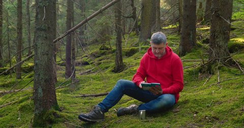 Man in nature. Outdoor travel concept.A bearded man is resting in a beautiful forest. He sits on the moss and reads a book. Slow motion. Ultra 4K