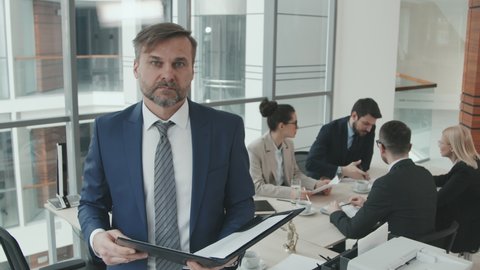 PAN medium slowmo portrait of mature male director or founder of law company posing for camera holding folder with legal documents with team of lawyers discussing case at meeting table in background