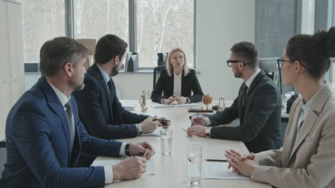 Medium shot of mid-adult blonde female director of law firm sitting at head by conference table talking to team of successful lawyers during meeting in office
