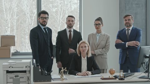 Medium PAN slowmo portrait of group of successful lawyers in formalwear posing for camera at workplace with their female leader sitting by desk in modern office room in big law firm