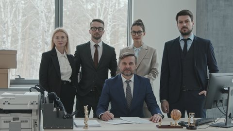 Medium slowmo portrait of group of five confident male and female lawyers in suits and formalwear posing for camera at workplace in modern office