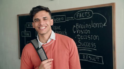 Young happy smiling confident Indian latin Hispanic high school college university student standing in classroom on blackboard background holding backpack on shoulder looking at camera. Portrait.