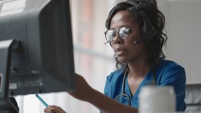 African Female medical assistant wears white coat, headset video calling distant patient on computer. Doctor talking to client using virtual chat computer app. Telemedicine, remote healthcare 