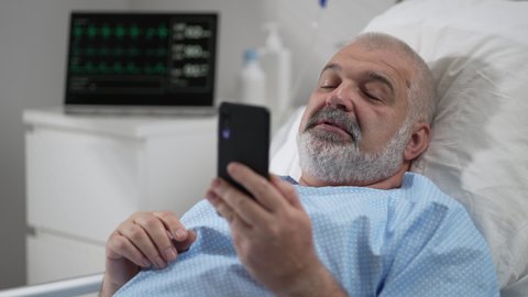 Hospital Ward: senior man Resting in Bed uses Smartphone for Video Call Conference Talk with Family and Friends. video call in hospital room
