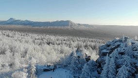 Aerial view of snowy winter forest with mountains in the background. Pine and spruce trees are covered with winter snow. Frosty sunrise or sunset. Tourists walk and admire the peaks of mountain range