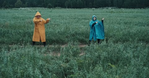 Man in yellow raincoat and a woman in blue raincoat dance in a field. They simultaneously point their index fingers in different directions. Opening and editing for video clips. Concept of joy and fun