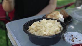 This close up video shows a carry out container of macaroni and cheese being scooped and served at park picnic table.