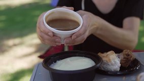 This close up video shows anonymous hands pouring brown gravy over mashed potatoes in slow motion on a sunny day at a park picnic table. 