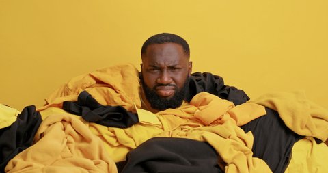 Unhappy dark skinned man frowns face from dissatisfaction looks around covered in heap of yellow and black clothes engaged in spring cleaning does daily domestic routines. Human drowned in lundry