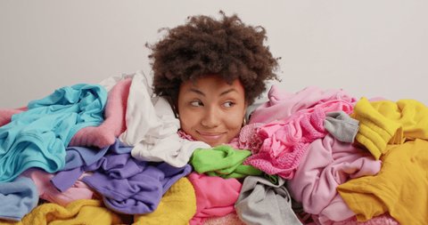 Curly haired woman buried in heap of laundry sorts out clothes in wardrobe has real chaos at home poses against white background. Afro American female covered with clothing stack for donation