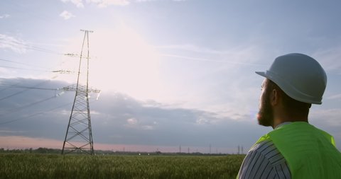 Engineer in yellow vest and helmet looks at tower with power transmission lines in spring evening backside view slow motion