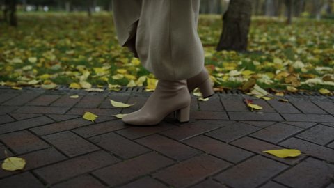 Close up woman legs in boots walking at walkway in autumn daytime. High heeled female shoes going slowly in beautiful fall season. Unrecognizable lady relaxing in october park.