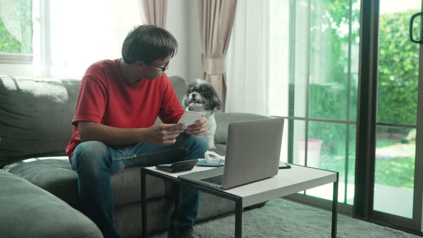 Asian man wear glasses red shirt and jeans with money and a calculator checks bills, calculates expenses, studies the credit balance sitting at the table at with a dog home,Concetp lifestlye finance.
