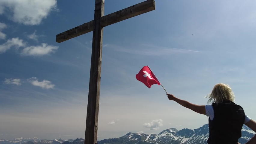 Woman with Swiss flag on top cross of the Piz Scalottas. Plessur Alps in Switzerland in Graubunden canton. Tourist resort for ski activities in winter and mountain bike in summer. SLOW MOTION