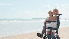 Elderly Asian woman sits in a wheelchair on the beach talking and smiling with her granddaughter.