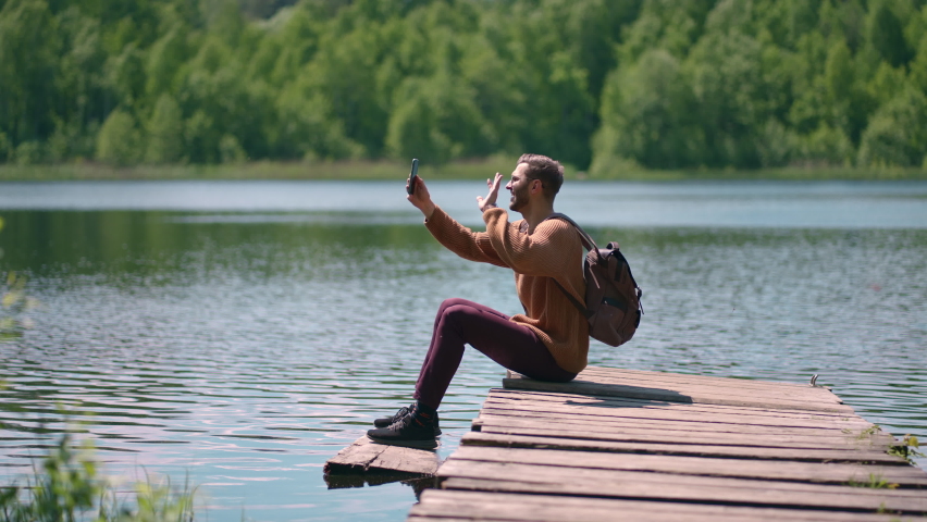 A man with a backpack sits on the dock of the lake and calls by video link on a mobile phone. Sitting on a lake in nature to call friends. Blogger shoots content for the channel