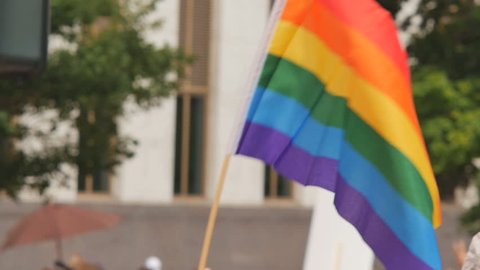 A rainbow flag waves as people walk by in the background with other rainbow flags and protest signs. slow motion. Symbol of LGBT GLBT transgender rights love equality 