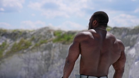 Muscular dark skinned sportsman outdoors. Bare chested male bodybuilder turning torso and walking among nature. Bodybuilding concept.