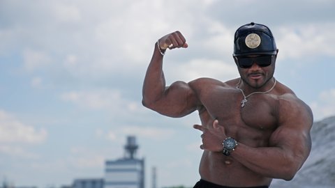 KYIV, UKRAINE - May 2021: Portrait of muscular athlete in the mountains. Shirtless african american sportsman in black cap and sunglasses showing his arms muscles outdoors.