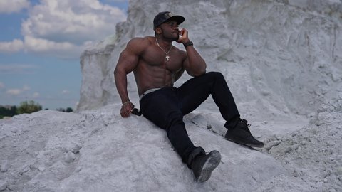 KYIV, UKRAINE - May 2021: Thoughtful black skinned guy sitting on white stone. Shirtless african american man with muscular body rests outdoors. Sportsman on hill in summer.