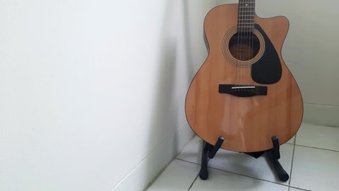 Bekasi, Indonesia - 09 July 2021: Putting guitar down to a guitar stand at the corner of a white room. Brown Acoustic Guitar with a black guitar strap.