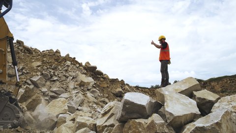 Zoom out view of foreman in protective uniform and respirator standing on heap of stones and gesturing thumbs up to excavator during work on quarry