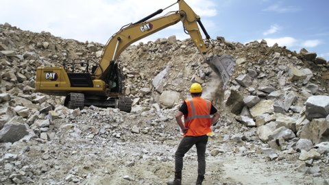 ZAPOROZHYE, UKRAINE - May 28, 2021: Zoom out view of man in hardhat and waistcoat holding hands on waist and watching excavator mining granite on quarry