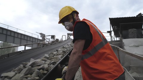 Low angle of man in protective uniform and respirator looking at conveyor belt with stone rubble and walking up path during work on quarry