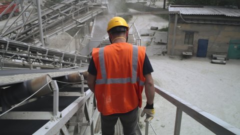 Tracking shot of anonymous man in hardhat and waistcoat walking on metal path and checking conveyor belt with rock rubble during work on quarry