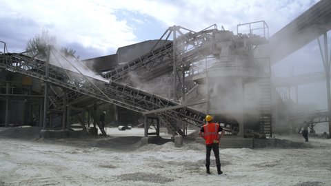 Zoom out view of anonymous man in hardhat and waistcoat looking at conveyor belts system during work in quarry on cloudy day