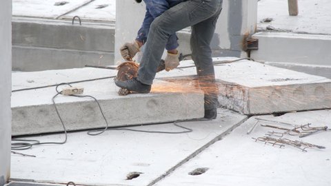 A professional builder cuts iron rebar with an angle grinder machine on a reinforced concrete slab at a construction site. Sparks fly from an electric circular saw. Construction of a new building.