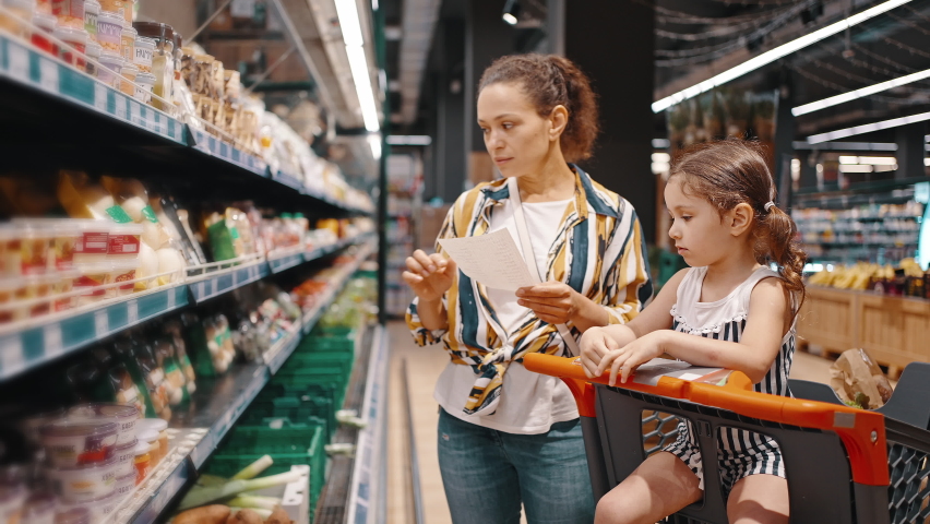 Woman takes packed products from the shelf, packed mushrooms and puts them in a trolley. Little girl and her mom, housewife, enjoy family shopping together. Royalty-Free Stock Footage #1075702784