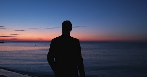 A silhouette of a man stands on the seashore in the early morning before the sun rises, a man looks towards the sunrise over the sea.
