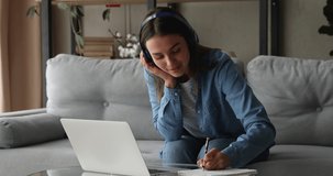 Focused millennial woman in wireless headphones looking at laptop screen, watching educational online lecture or attending distant class via video call, writing notes, communicating with teacher.